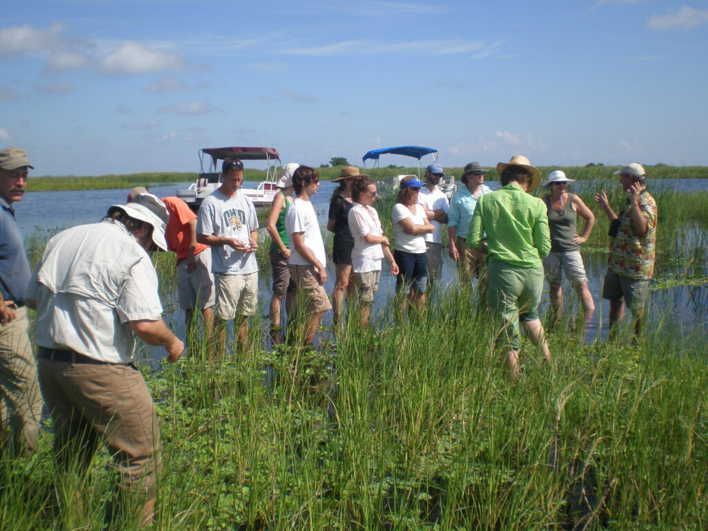 A group of students and faculty at a water body listening to a speaker.